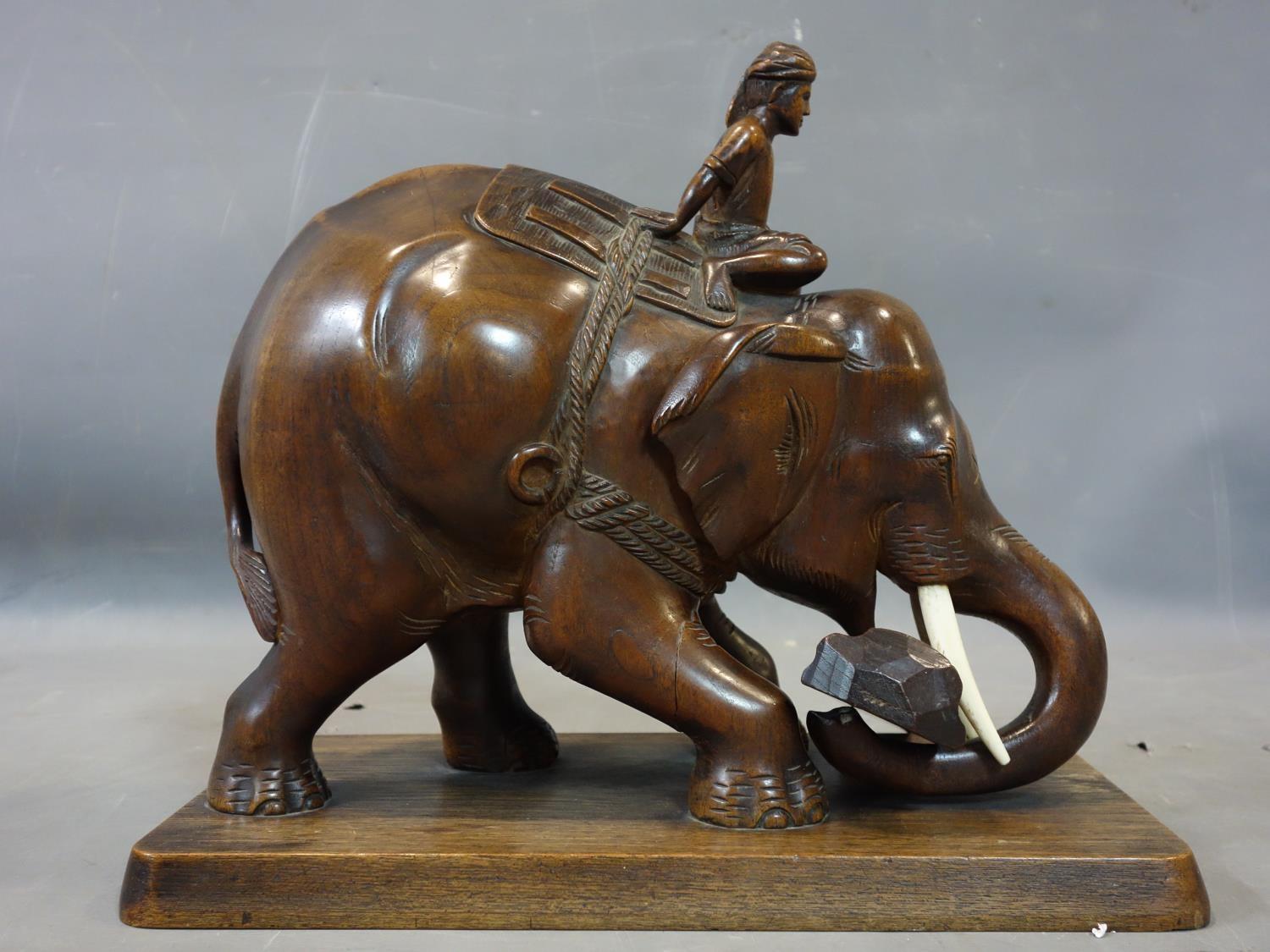 A teak carving of a man riding an elephant picking up a log, with bone tusks, on rectangular teak