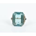 A gem-set ring centred with a blue-green emerald-cut synthetic gemstone, each shoulder highlighted