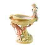 A Royal Dux early 20thC porcelain figural comport, modelled with two maidens, one sitting atop the