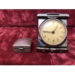 A small eight day travelling clock in a folding case together with a small silver pill pot.