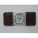 A small travel clock having brown leather slide action cover, folding stand and Roman numeral dial