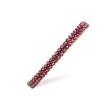 A GARNET BANGLE, of oval form with reeded clasp and safety chain, designed with two rows of fine-cut