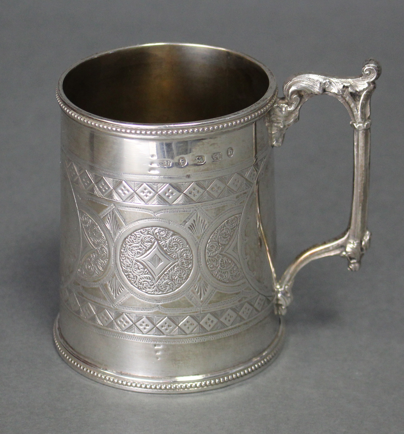 A Victorian silver round tapered mug with cast gothic leaf-scroll handle, beaded rims, & engraved