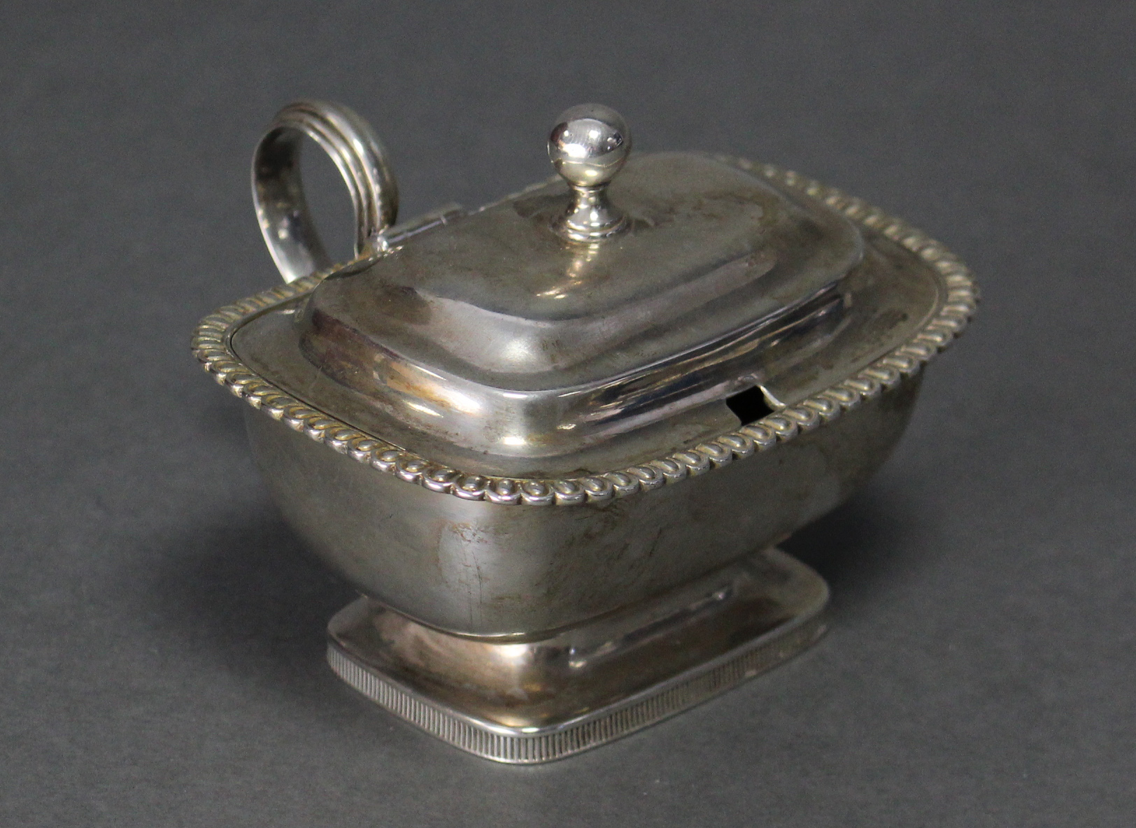 A George III silver mustard pot of compressed rectangular form, with gadrooned rim, ball finial to