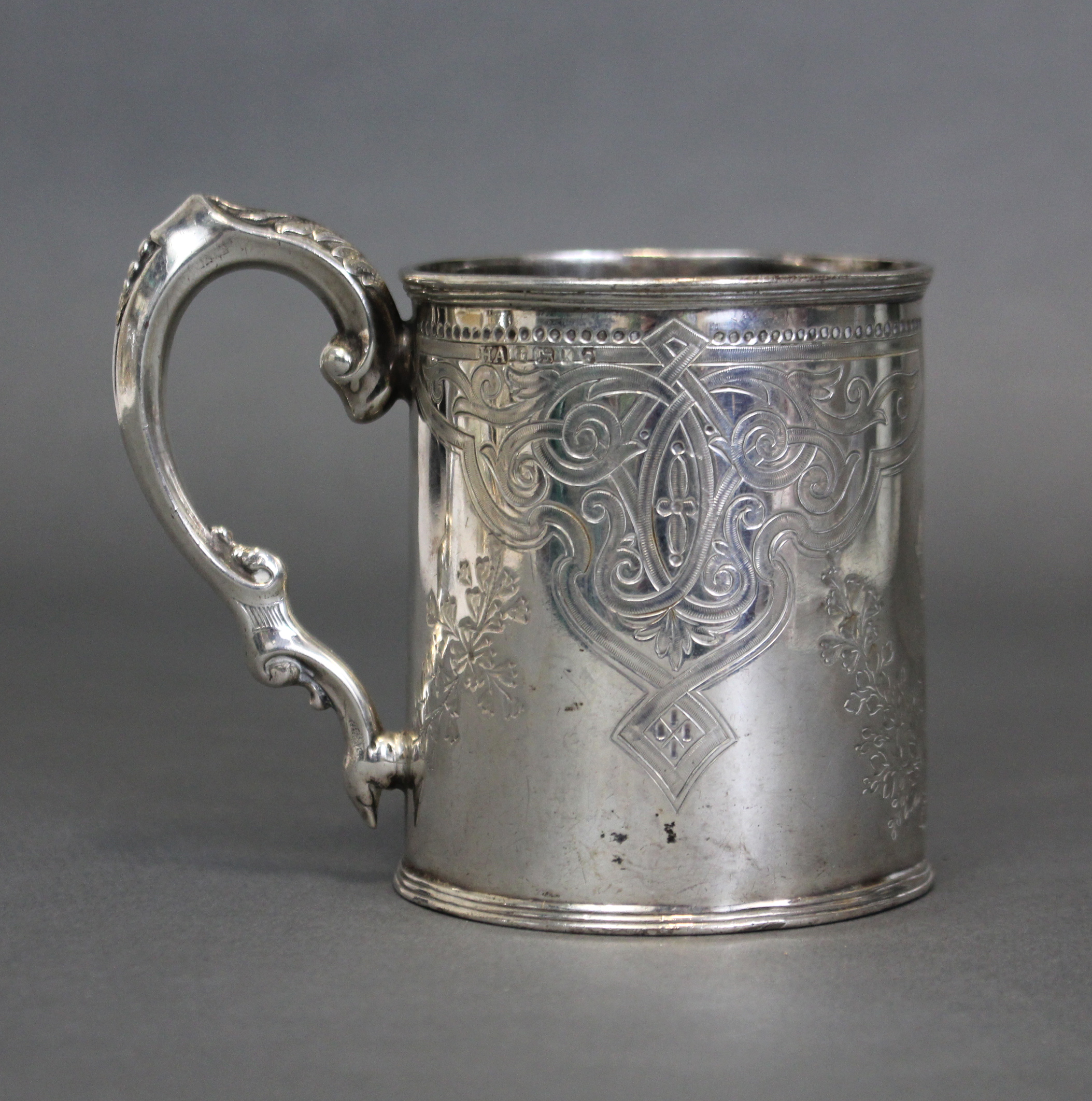 A Victorian silver cylindrical mug with engraved decoration, shaped scroll handle & reeded foot;