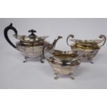 An Edwardian three piece silver tea set of oval multi-panelled bowl design, on C-scrolled pad feet,