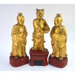 Three gilded wood Chinese figures; each one standing on a scarlet stand; the tallest 34 cm high,