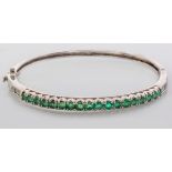 A 9CT WHITE GOLD AND EMERALD BANGLE, the top half claw-set with eighteen emeralds and flanked by