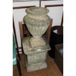 A WELL WEATHERED CAST CONCRETE GARDEN URN standing on a cast concrete pedestal plinth, decorated