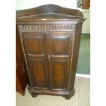 AN OAK FINISHED FREE-STANDING CORNER CUPBOARD with carved frieze over twin cupboard doors, supported