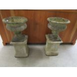 A pair of weathered composition stone urns, on relief decorated pedestals