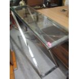 A modern glass topped side table having a folding chrome cross frame, 31" h x 51"w