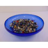 A blue studio glass dish with a quantity of glass decorative pebbles and glass sweets