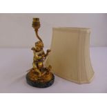 A gilded metal table lamp in the form of a putti on raised oval marble base to include silk shade