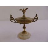 A brass tazza of classical form with bird finial, scroll handles on raised circular marble stand