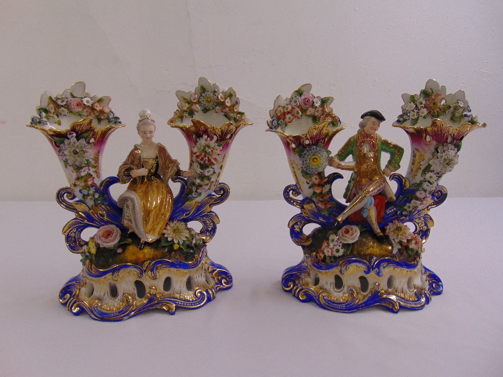 A pair of ceramic figurines in 18th century costume flanked by florally encrusted vases