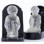 A pair of black glass book ends surmounted by moulded opaque glass figures of seated children, by