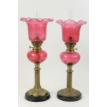 Two similar cranberry glass and brass pedestal oil lamps, each having a moulded bell shaped shade