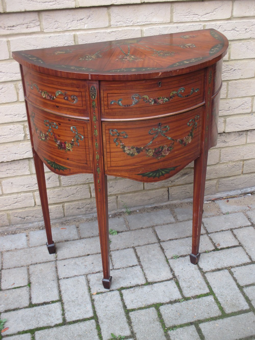 An Edwardian painted satinwood demi-lune cabinet, decorated with floral swags to the hinged top