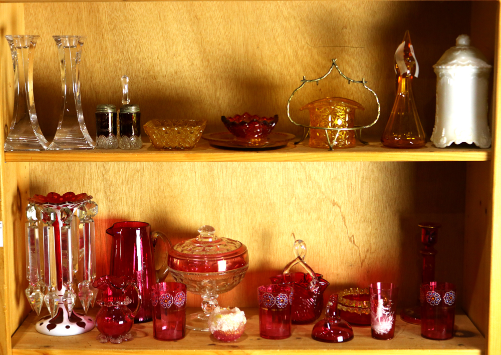 Two shelves of mostly cranberry glass, including a Mary Gregory tumbler, together with a lustre,