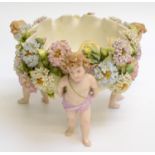 A porcelain bowl supported by three cherubs, the interior floral enamelled, the rim crimped,
