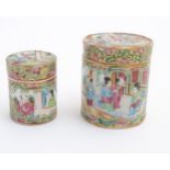 Two Cantonese famille rose cylindrical lidded pots with hand painted panelled decoration depicting