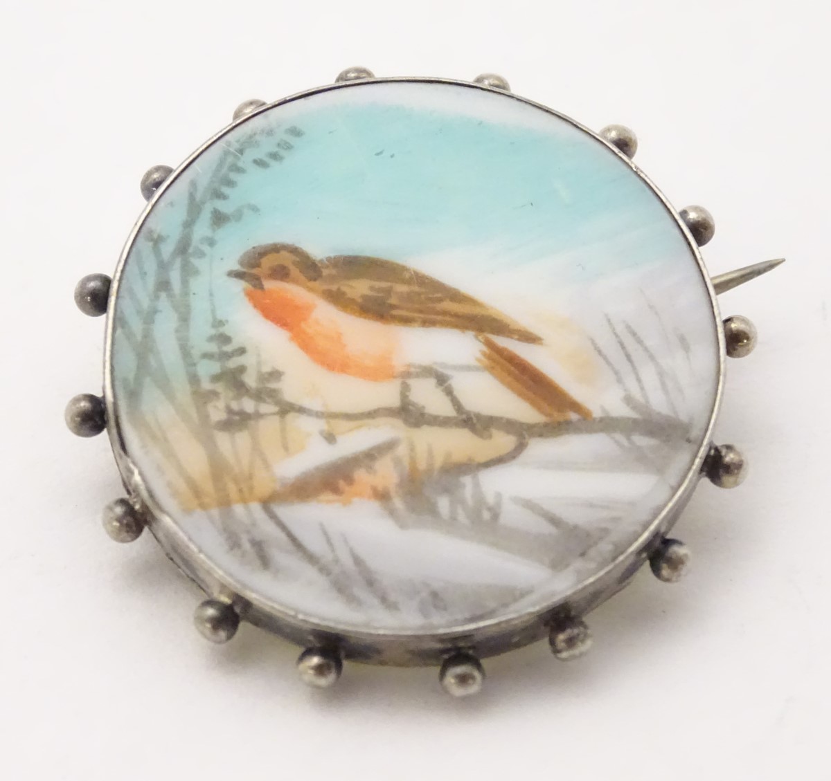 A white metal brooch with hand painted scene to centre depicting a robin in a snowy landscape.