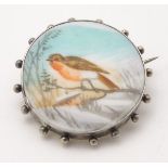 A white metal brooch with hand painted scene to centre depicting a robin in a snowy landscape.
