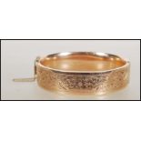 A 9ct gold bangle. The bangle having foliate decoration and hidden clip clasp with safety chain.