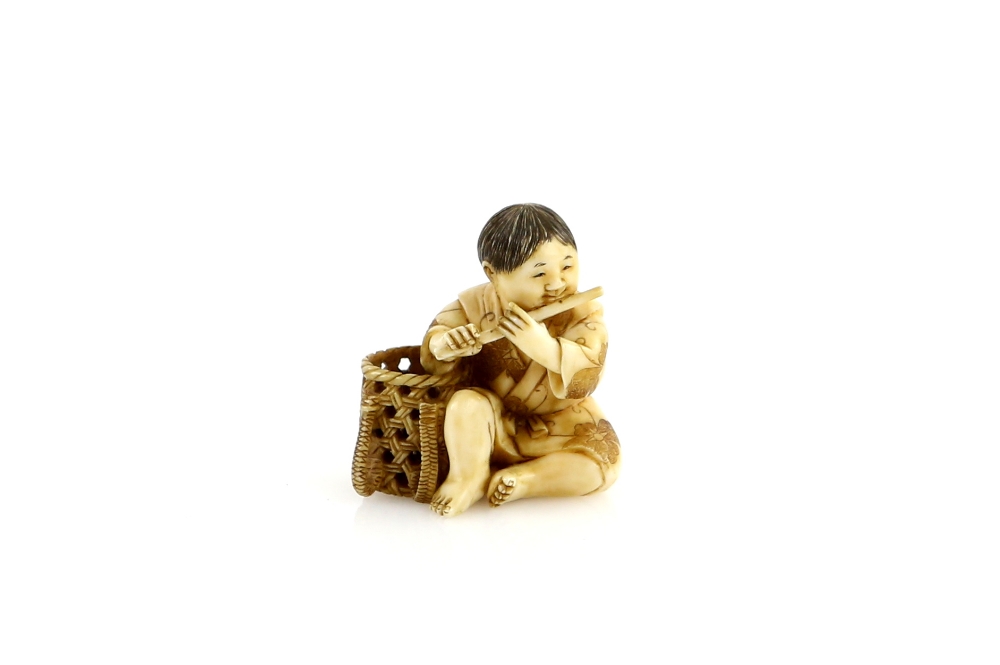 A stained ivory netsuke of a boy flautist, seated beside a basket, signed YoshimitsuProvenance: