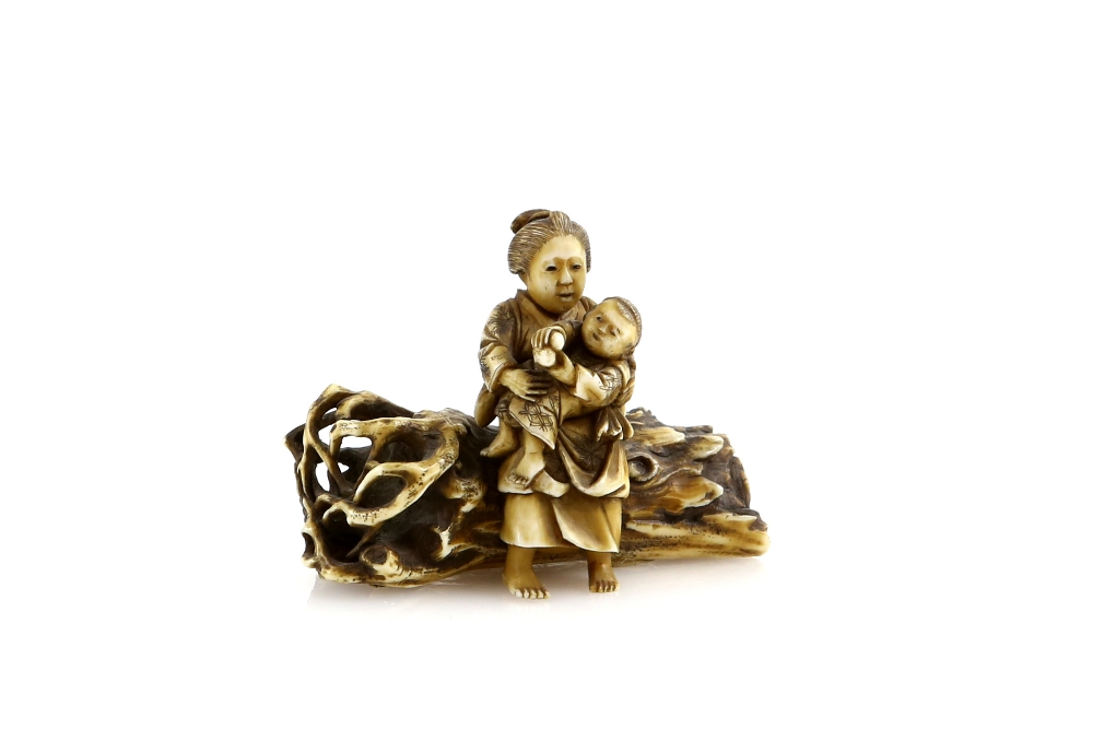 A small stained ivory netsuke or okimono of a mother, seated on a log with her child; two kanji on