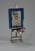 An Italian post 1968 800 white metal, enamel and glass model of an easel, with pallet and