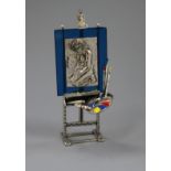 An Italian post 1968 800 white metal, enamel and glass model of an easel, with pallet and