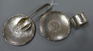 Two white metal dishes each inset with Chinese coin, a pair of Chinese sugar tongs and a napkin
