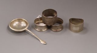 Four silver serviette rings and a silver tea strainer.