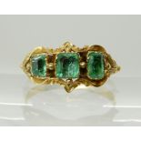 A BRIGHT YELLOW METAL VICTORIAN THREE EMERALD RING in scrolled foliate design mount, largest emerald
