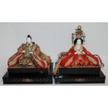 A PAIR OF JAPANESE PAPIER MACHE DOLLS with detailed silk costume, seated on rattan and lacquered