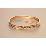 AN EDWARDIAN STIFF HINGED BANGLE, the open reeded bands tied by a knot and one section having bright