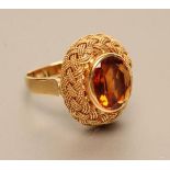 A MODERN 18CT GOLD DRESS RING, the oval facet cut topaz open back collet set to a plaited rope