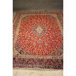 A KASHAN CARPET, the finely woven claret red field with navy blue spandrels and central gul and