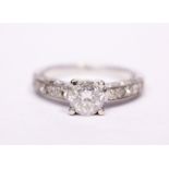A DIAMOND RING, with a central round cut diamond set in a four claw setting with diamond shoulders &
