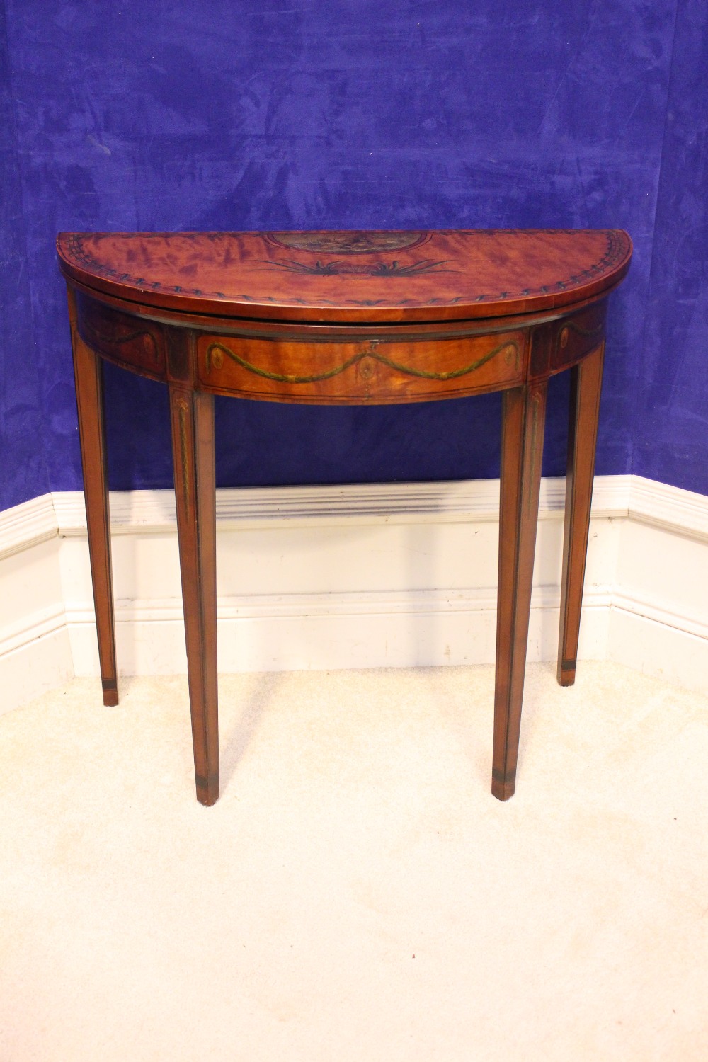 A DEMI LUNE SATINWOOD FOLD OVER CARD TABLE, decorated with hand painted motif of foliage and cherubs