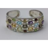 A gem set open-end bangle, claw set with twenty-three oval cut stones in silver, with a sprung