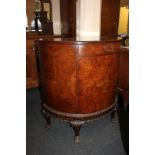 An early 20th century burr walnut demi-lune side cabinet with drawer above panel door, on carved