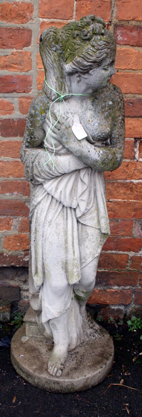 A weathered composite garden statue, modelled as a young woman with hands clasped to loose robes,