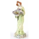 AN ART NOUVEAU NOVE FAIENCE FIGURINE OF A YOUNG LADY HOLDING A FLOWER decorated with polychrome