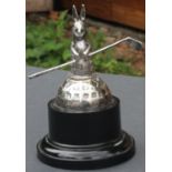 A silver golf trophy featuring a rabbit or hare holding a golf club and sat on a square mesh golf