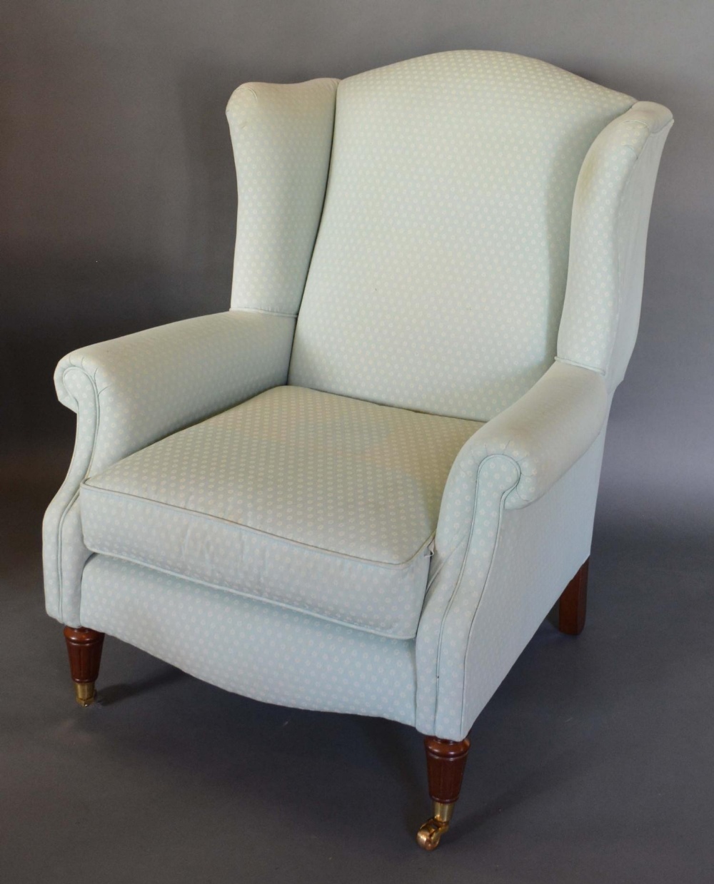 An Upholstered Wing Back Arm Chair with turned reeded legs, brass caps and castors