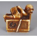 A SIGNED JAPANESE MEIJI PERIOD STAINED IVORY NETSUKE OF AN ONI CATCHER, lying atop a box from the