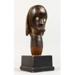 A CARVED TRIBAL HEAD with bone eyes on a square wooden base. 1ft 1ins high.